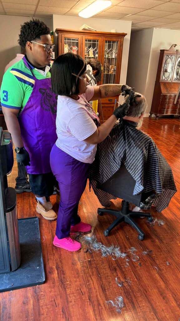 A woman cuts a man's hair while a student watches.