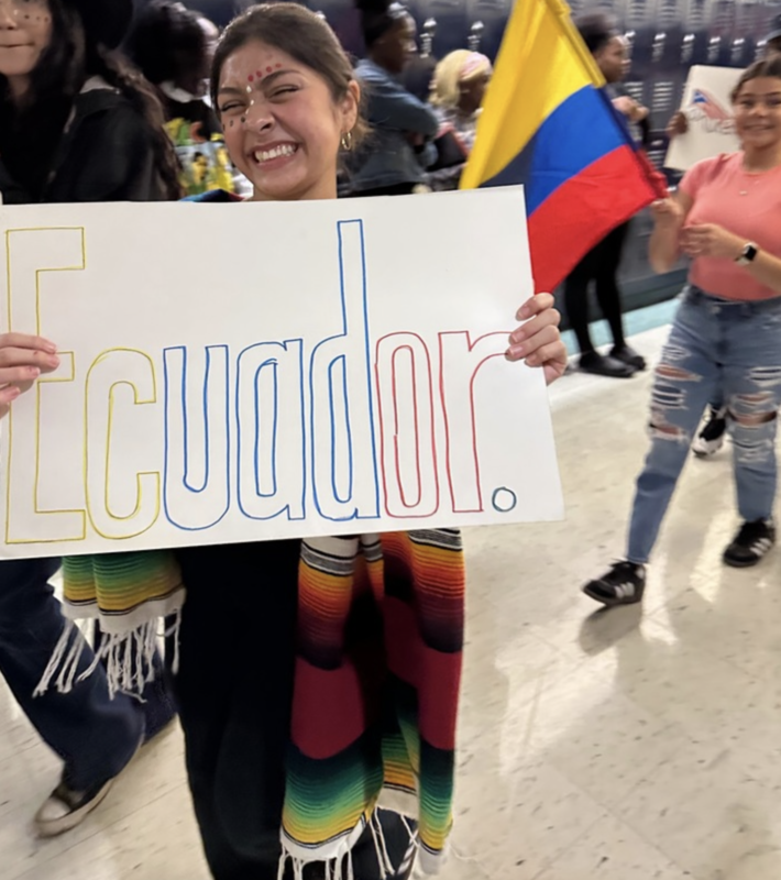 A student holds a sign that says "Ecuador." while standing in a school hallway. Behind her, several students stand behind her holding posters and flags.