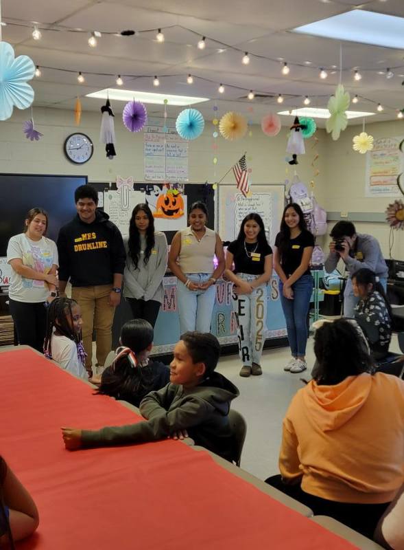 Former students speak to a class of current students at an elementary school.