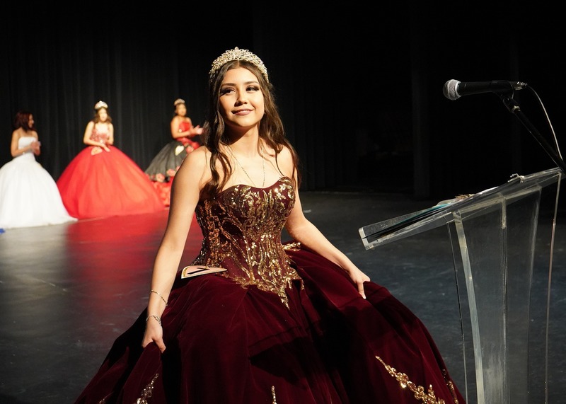 A girl with a gown and a tiara on holds her dress while standing in front of a podium with a microphone attached to it.