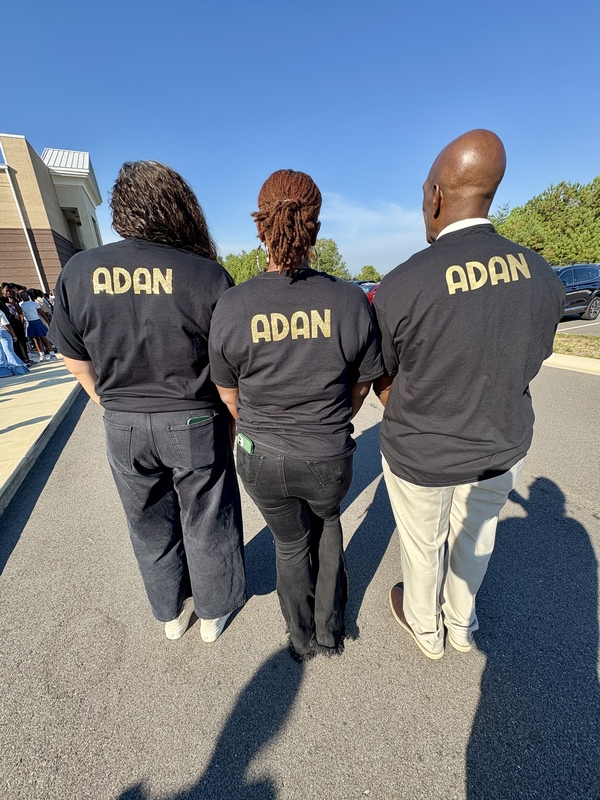 The photo is of three adults standing outside a school. Their backs face the camera, and their shirts say "Adan" on the back.