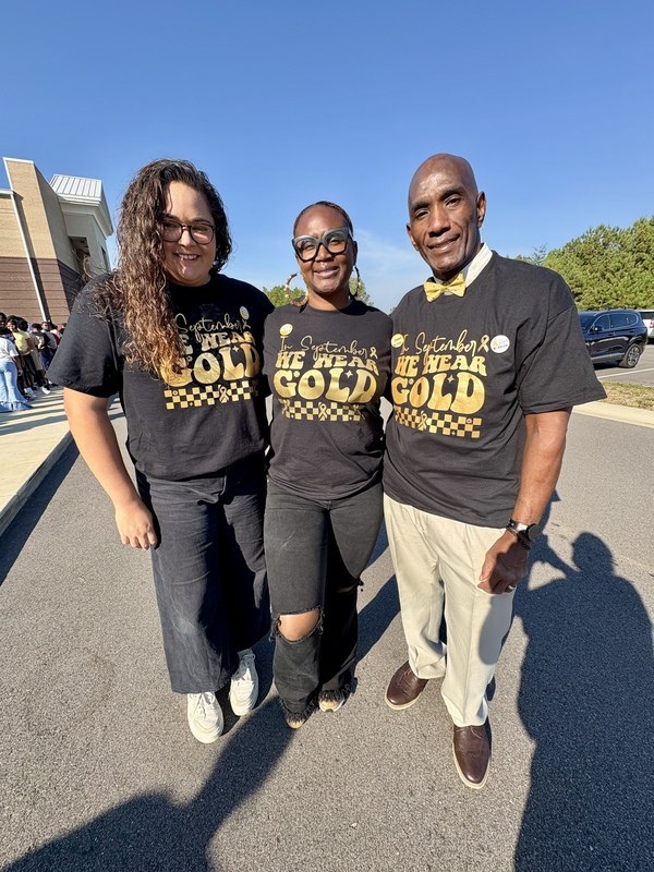 A group of adults stand together, smiling. They have on shirts that say "In September, we wear Gold."