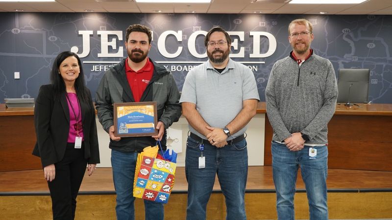 A woman and three men stand together in a board room and smile. One of them is holding an award and a gift bag.