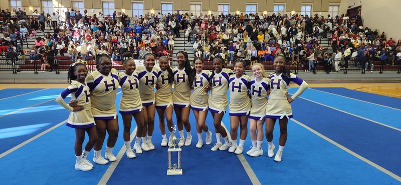 Hueytown Middle School cheerleaders pose for a photo with a trophy while standing on a cheer mat in a gym.