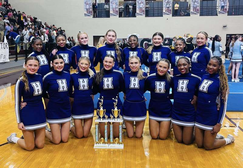 Hueytown High School cheerleaders pose with two trophies while standing and sitting in a school gym.