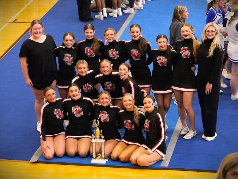 Oak Grove cheerleaders stand and sit while smiling and posing with a trophy inside a school gym.