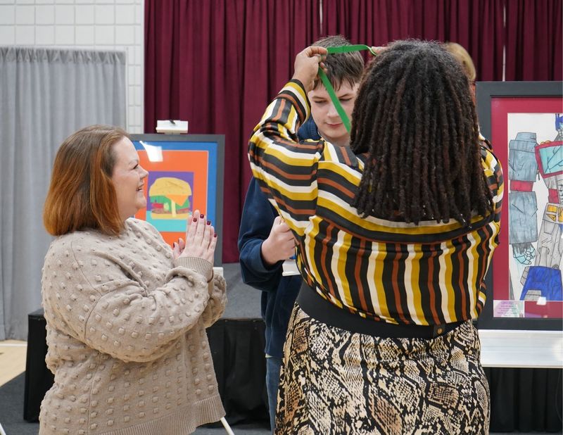 A student receives a medal at The Exceptional Art Show. A woman stands next to him and claps.