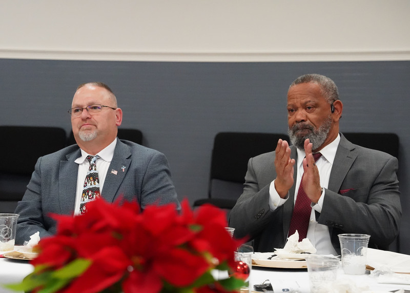 Two men sit at a table during a meeting. The man on the left is listening and the man on the right is clapping. 