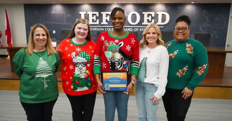 JEFCOED finance employees stand together for a photo inside the board room and smile. Each of them wear holiday sweaters. The employee in the middle, Monica Spencer, holds her JEFCOED Strong award.