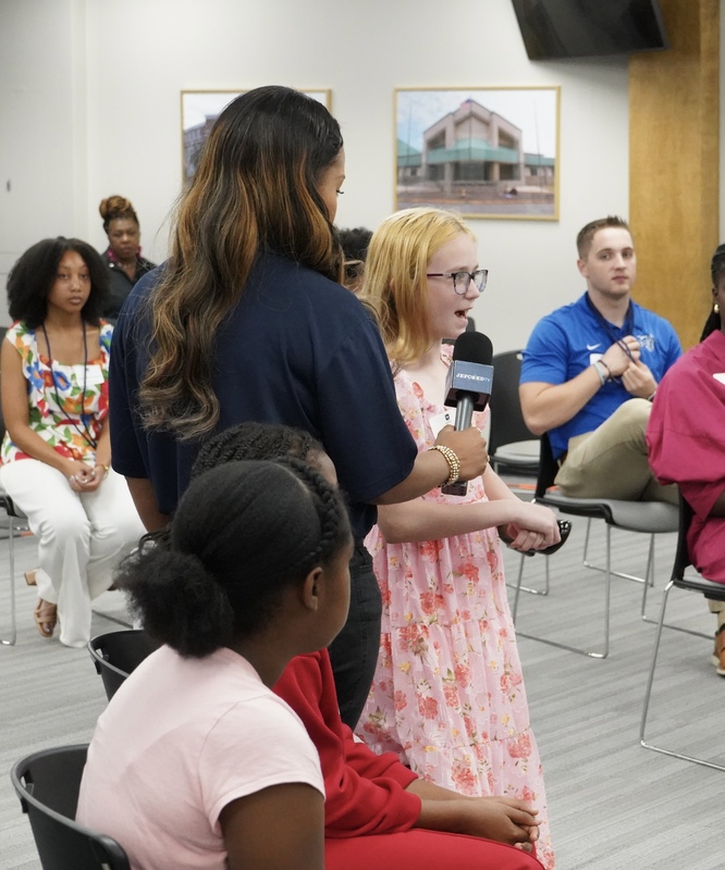 A woman holds a microphone for a student to speak into. They stand in the JEFCOED board room. Students sit in chairs around them.