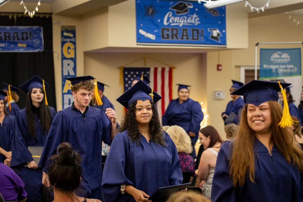 Graduates of the Adult Ed program in their caps and gowns with "Congrats Grad!" signs behind them at the Corner Connection graduation ceremony.