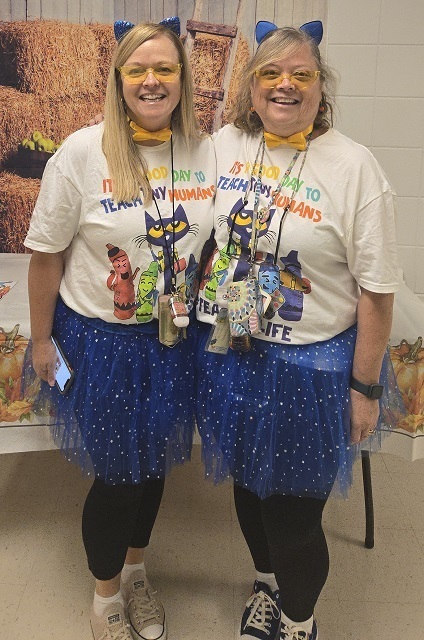 Two women dressed as cats for a school event, wearing blue cat ears, yellow glasses, matching shirts that read "It's a good day to teach tiny humans," and blue polka dot tutus. They stand side by side, smiling at the camera, in front of a harvest-themed background.
