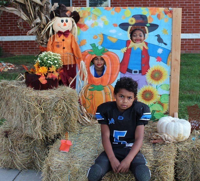 Three children posing in front of a fall-themed photo booth setup with hay bales and pumpkins. Two children peek through cutouts shaped like a pumpkin and a scarecrow in the background, while one child, dressed in a black football uniform, sits in front looking serious.