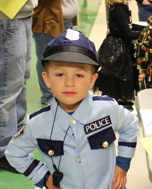 A young child dressed as a police officer with a light blue uniform, badge, and cap. He stands with his hands on his hips, looking confidently at the camera in a school hallway.