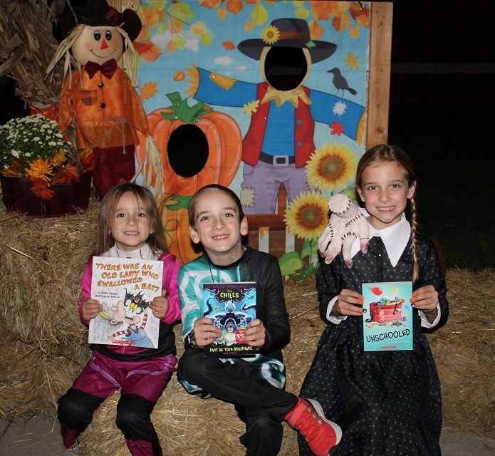 Three students sitting on a hale bale in front of a fall photo display holding up books.