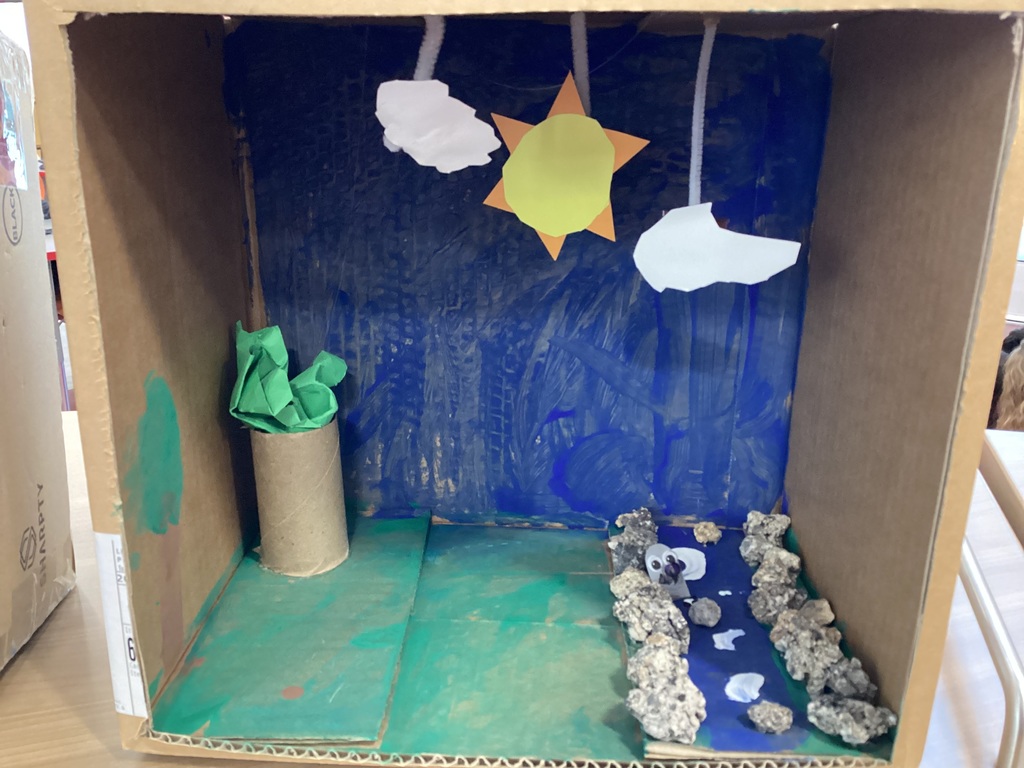 A box painted green for grass, blue for the sky, with a sun and clouds cut out of paper.