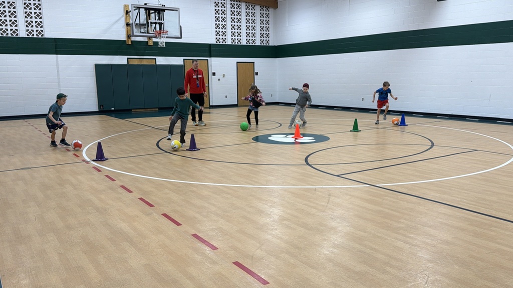 Students dribble around cones competing in a relay race, while a teacher provides feedback in the background.