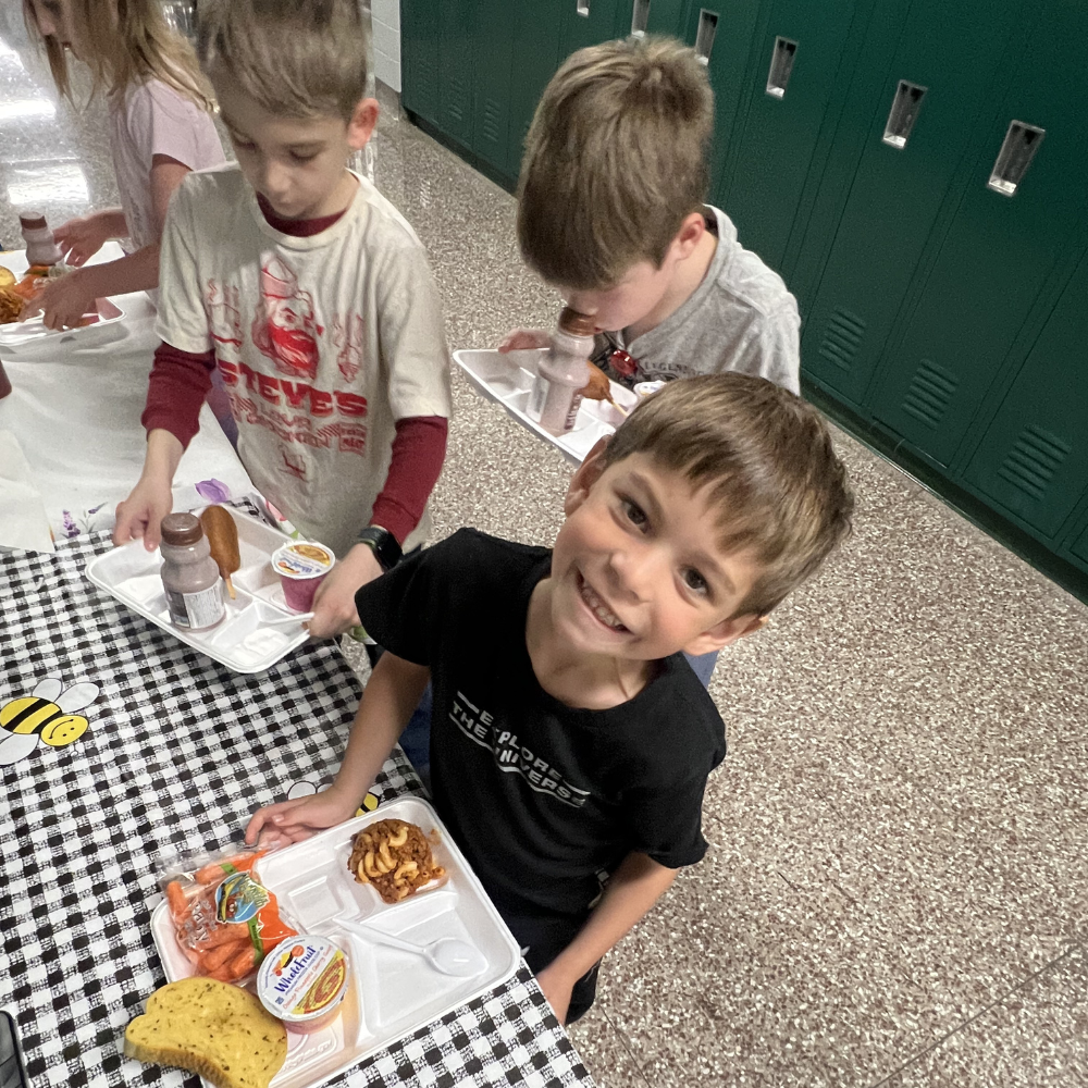 An elementary-aged boy smiles up at the camera with his lunch tray.