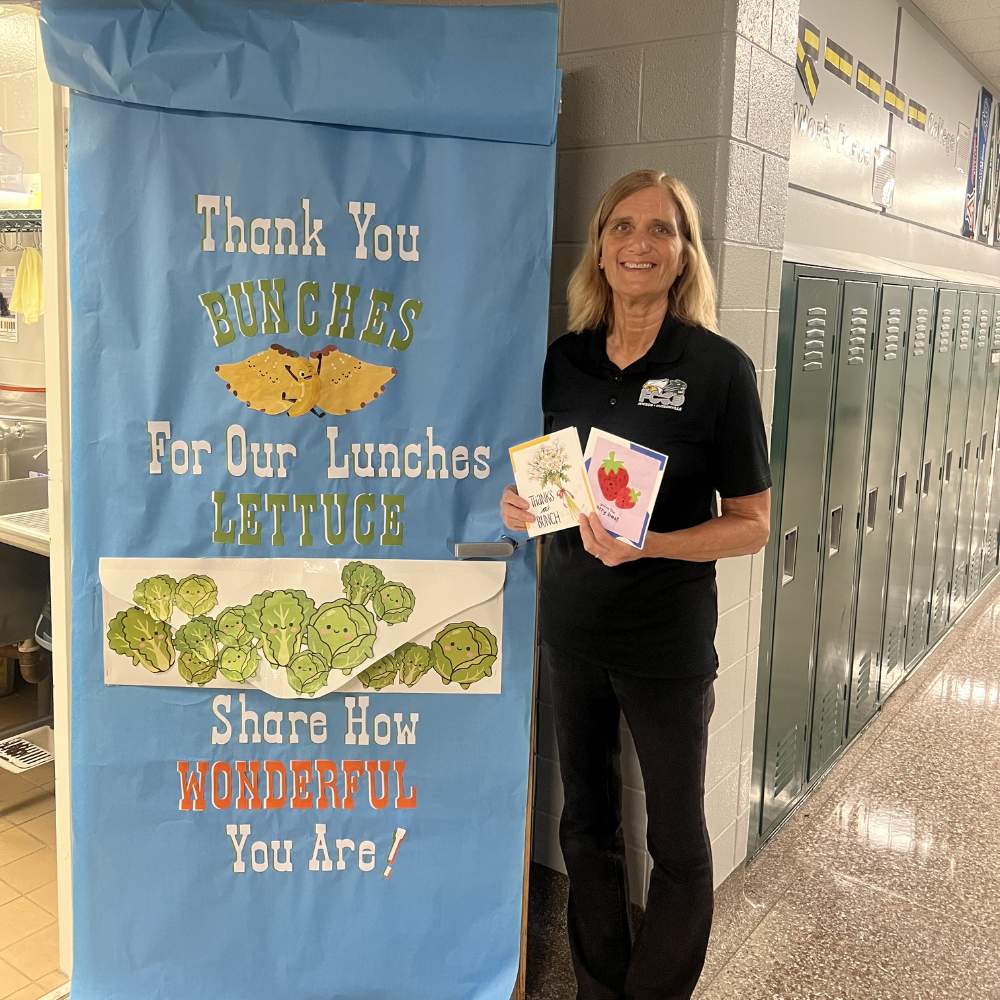 Ms. Jan stands in front of the kitchen door with artwork that reads "Thank you bunches for our lunches! Lettuce share how wonderful you are!" holding student-made cards.