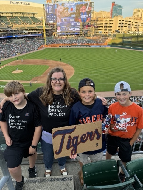 A teacher and three male students pose at Comerica Park with the baseball field and scoreboard in the background.