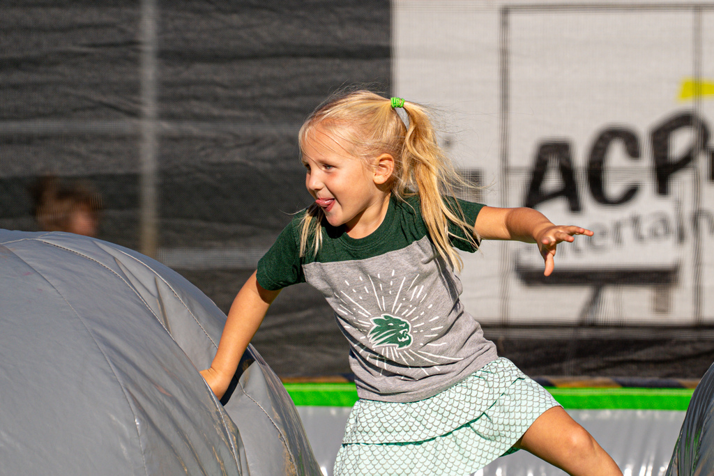 A young girl with pigtails laughs as she jumps into a large inflatable structure, background shows a sign.