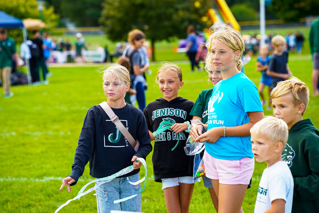 Kids stand in a grassy area. Some wear backpacks. A girl holds green ribbons. Behind them, more children and adults are present.