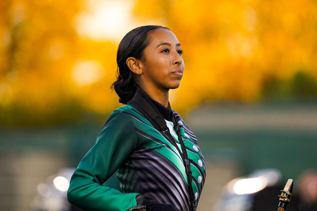 A woman in a green and black uniform stands facing left. Blurred autumn trees are in the background.