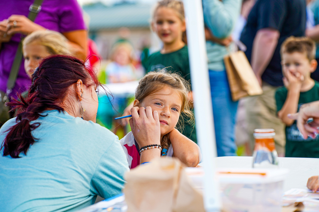 A woman applies face paint to a child's cheek at an outdoor event. Behind them, other children and adults are present.