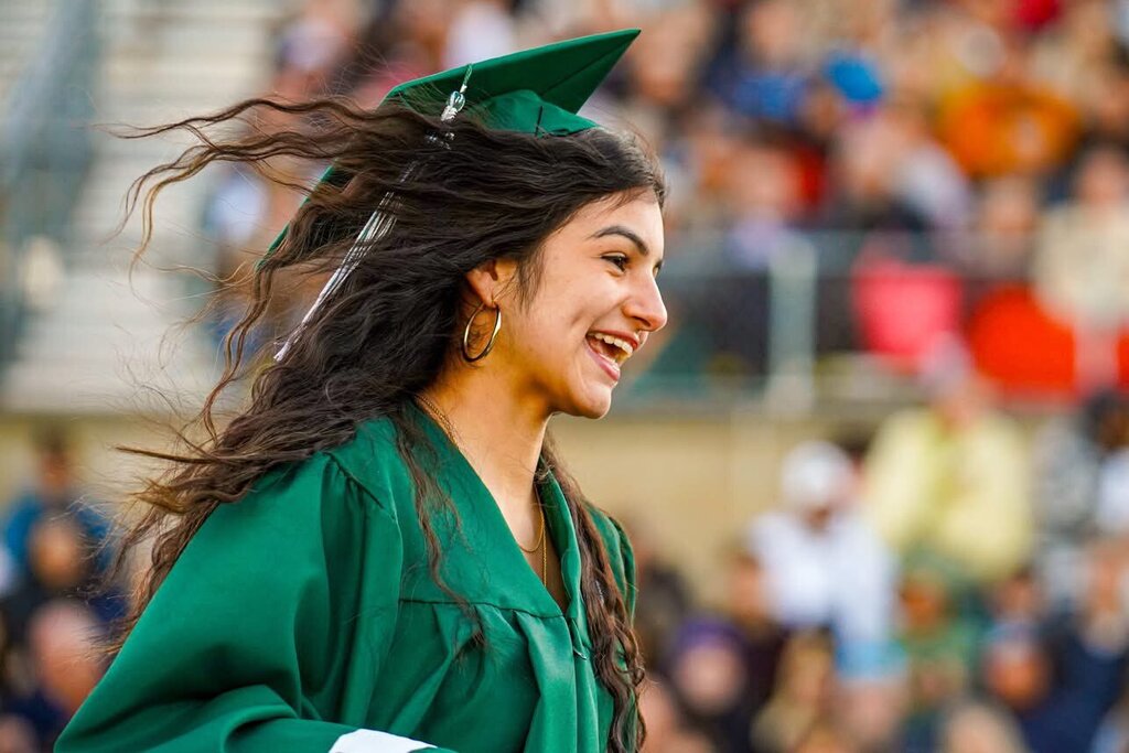 A woman in green graduation garb, cap, and earrings walks in front of a crowd.