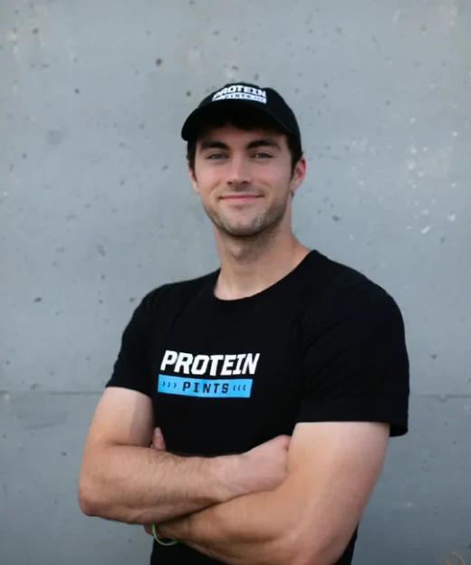 Man in black cap and shirt standing with arms crossed against a gray wall.