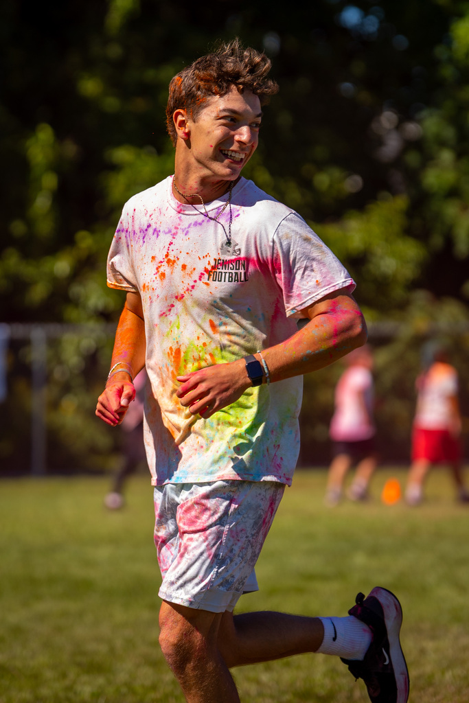 A young person runs on a grassy field, wearing a white shirt and shorts, both covered in multicolored paint.