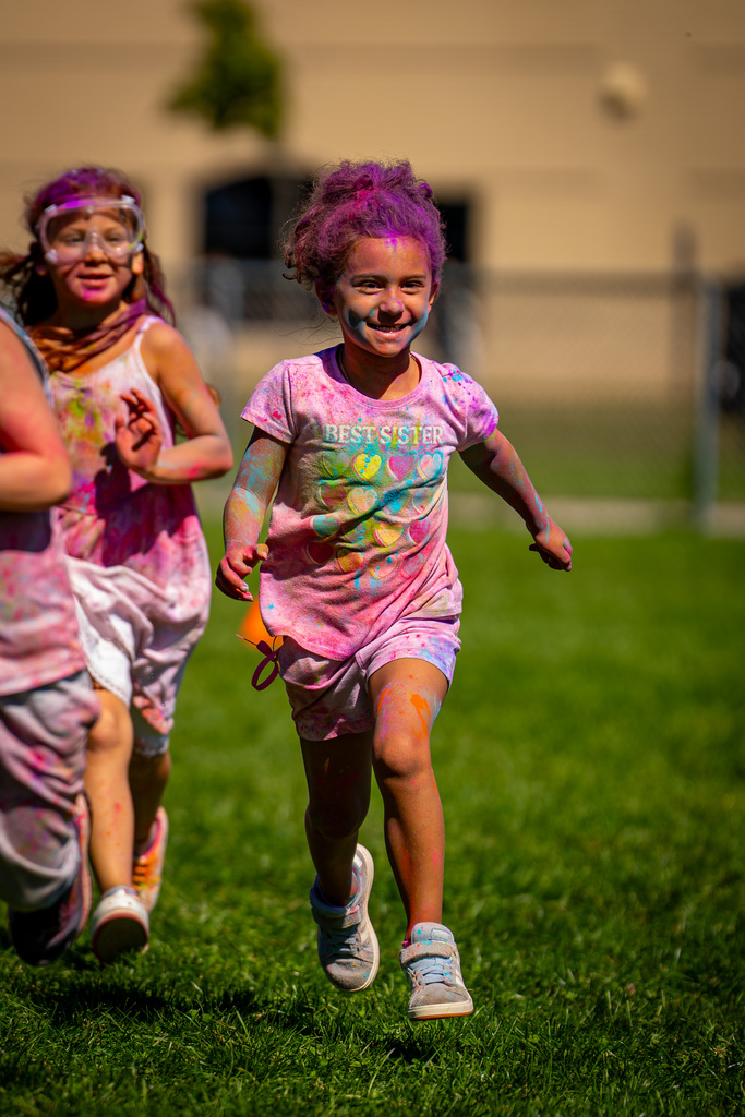 Two children run on grass. One wears a pink shirt, and the other wears a purple wig. Both have colorful paint on their faces.