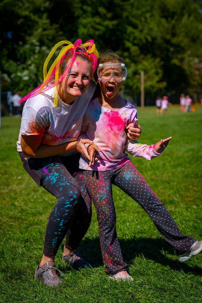 Two children covered in color stand on a grassy field. They smile and look at the camera.