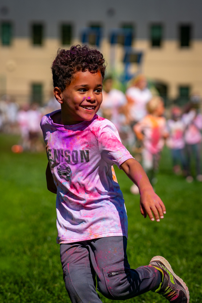 A young boy in a pink and white tie-dye shirt, running on green grass, with a large building in the background.