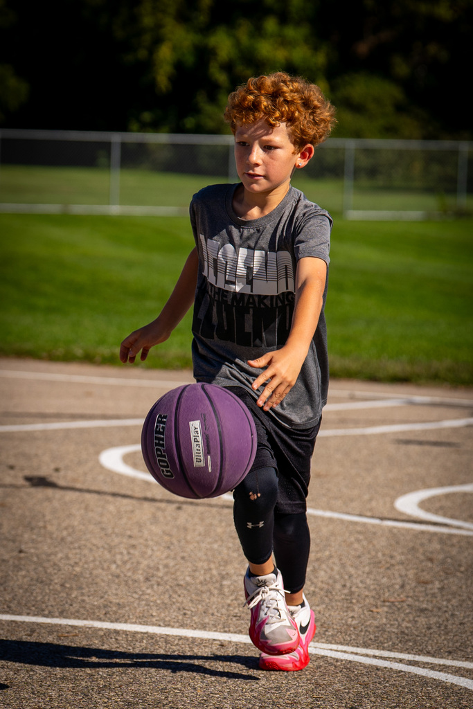 A young boy in a gray shirt, black shorts, and pink shoes dribbles a purple basketball on an asphalt court.