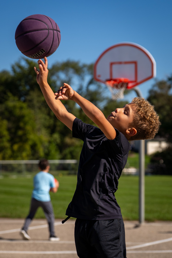 A child reaches for a ball over a basketball hoop. A second child stands in the background.