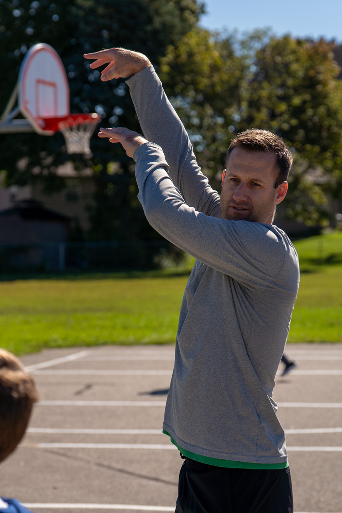 A man in gray long sleeves throws a ball at a basketball hoop. Trees and a house are visible in the background.