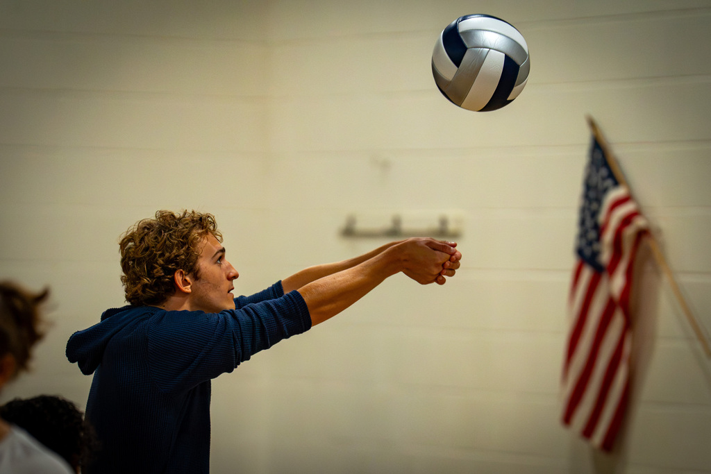 A person serves a volleyball in a gym. A flag is nearby. A wall is in the background.