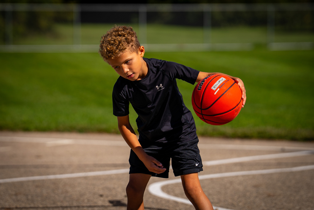 A child in a black shirt and shorts dribbles a basketball on a basketball court. Green grass and a fence are in the background.