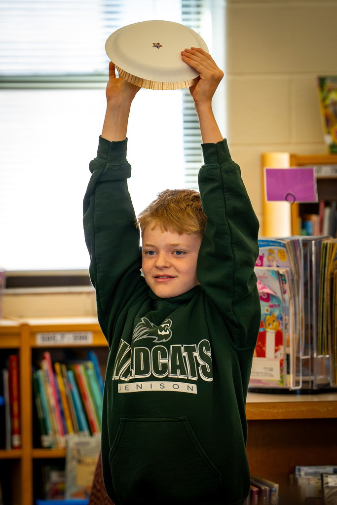 A child in a green sweatshirt raises a large paper plate above his head indoors near a bookshelf.