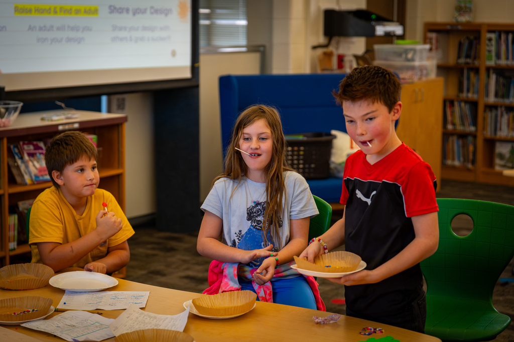 Children in a classroom, one holding a pie, others smiling. A whiteboard is in the background.