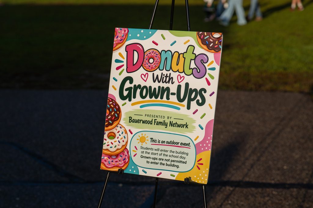 Brightly colored sign with "Donuts with Grown-Ups" in large letters. Colorful donuts on the sign. Background shows a grassy area.