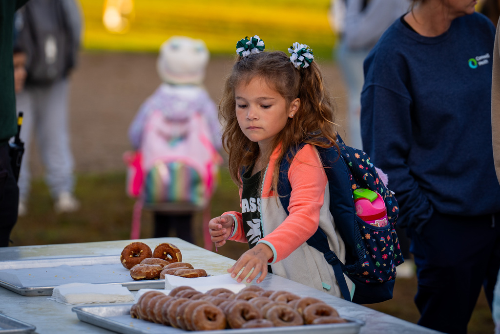 Child wearing backpack reaching for doughnuts at table; another child and adults are in the background.