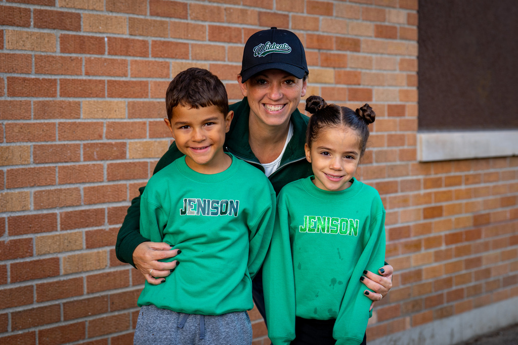 Two children and a woman in green shirts pose against a brick wall. The woman wears a black cap.