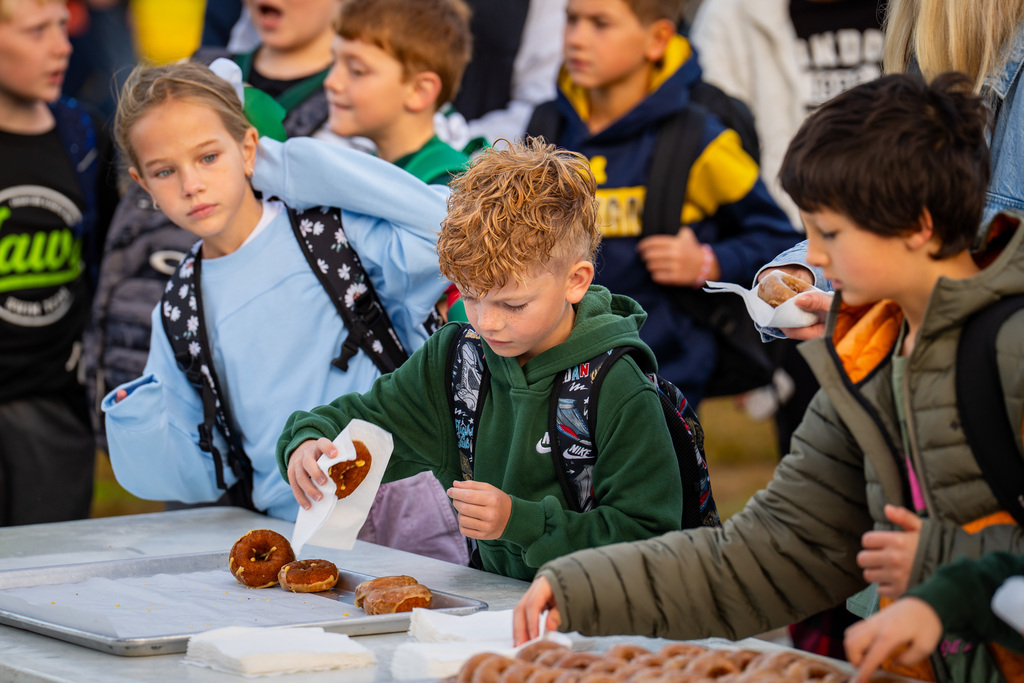 Group of children at an event, some eating donuts at a table, others standing nearby, with blurry background.