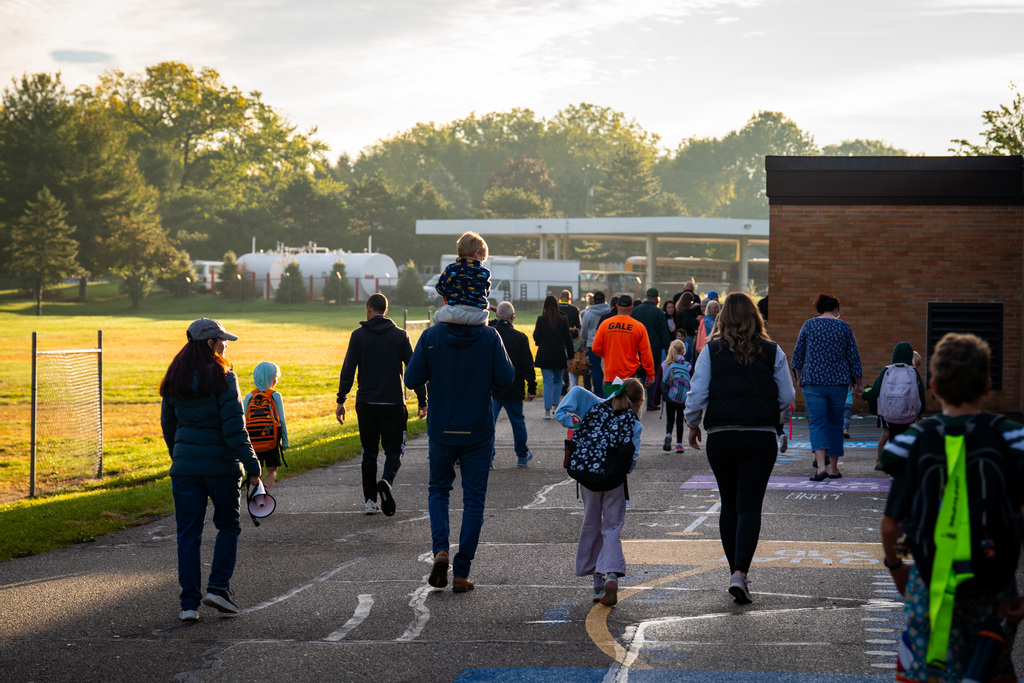 Group of people walking on a sidewalk at sunrise. Background includes a fence, trees, and a building.