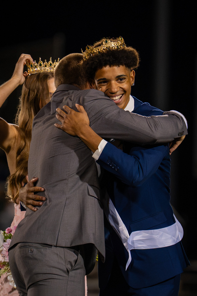 Three people on a field, two in suits and ties, one wearing a crown, hugging and smiling. Homecoming king and queen crowned.
