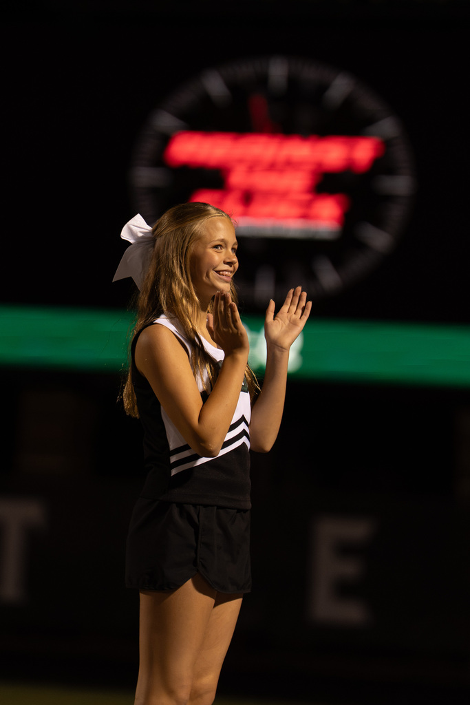 A cheerleader in a black and white uniform stands on a field, clapping hands, with a large clock in the background.