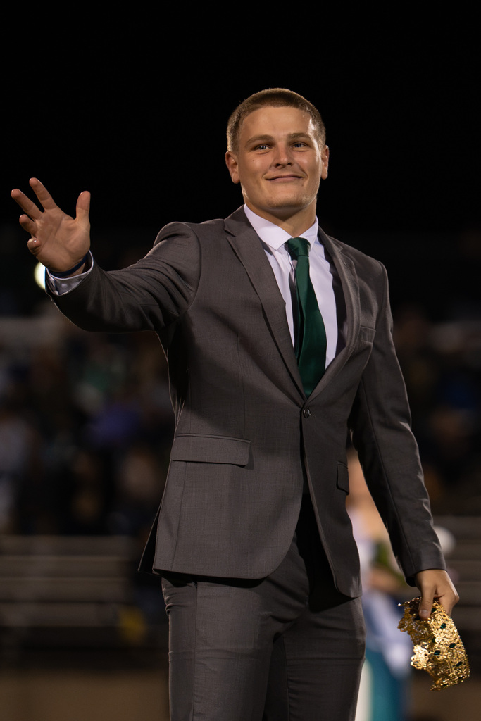 A man in a gray suit and green tie waves while holding a trophy. Background shows bleachers.