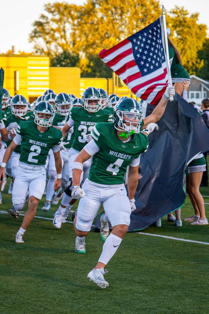 Football players in green uniforms running across the field, one holding a flag and another carrying a black flag.